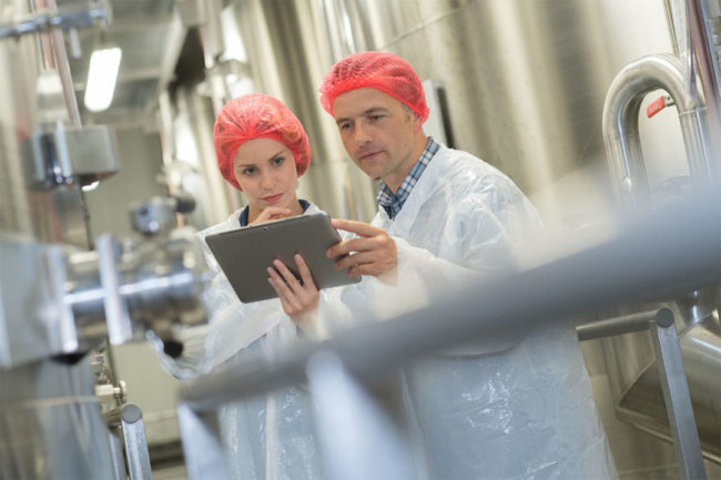 Man and woman wearing red hair nets and white laboratory coats study information on a tablet.